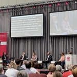 Ein Podium wo 6 Personen drauf sitzen, Köpfe von Menschen im Publikum unten im Vordergrund. 2 Großleinwände zeigen das gesprochene Wort sowie einzelne Gäste vom Podium in Nahaufnahme.
