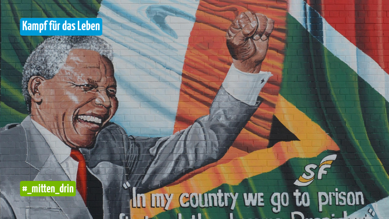 Wandgemälde von Nelson Mandela mit erhobener Faust vor der südafrikanischen Flagge. Dazu stehen die Texte „Kampf für das Leben“, „#_mitten_drin“