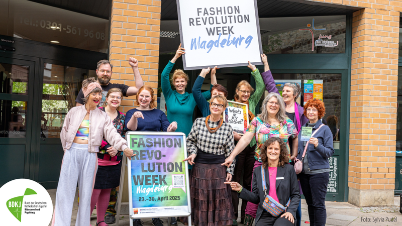 Eine Gruppe von freudigen Menschen, die ein großes weißes Schild hochhalten. Auf diesem Schild steht Fashion Revolution Magdeburg geschrieben.