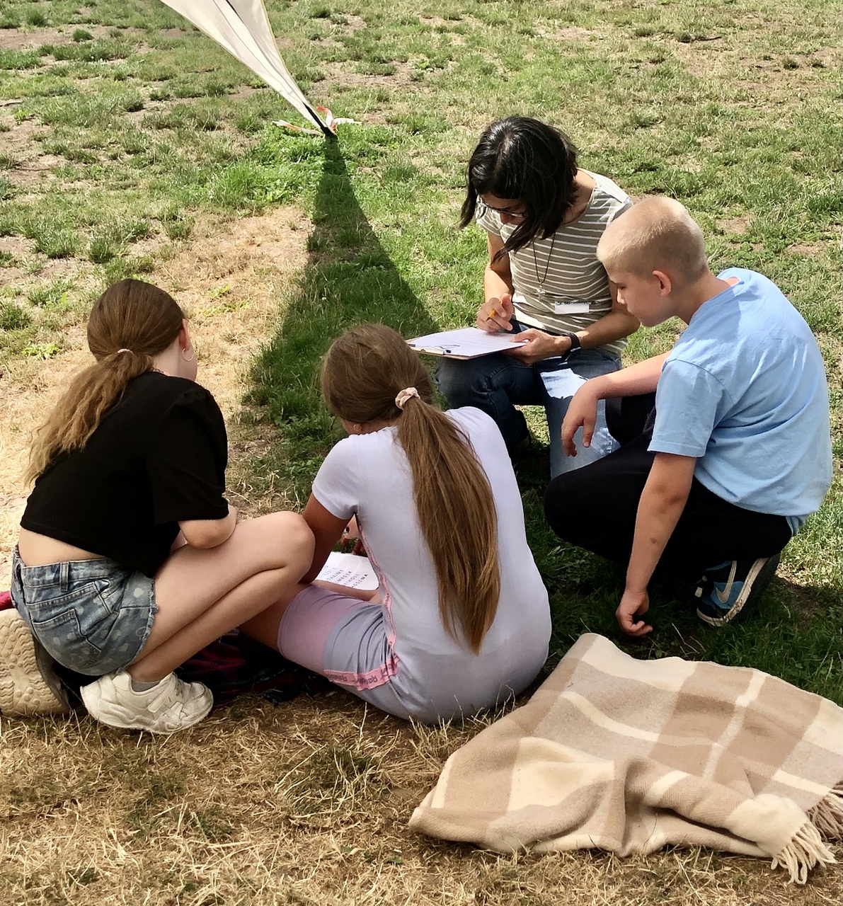 Zwei Mädchen, ein Junge und eine Frau sitzen bzw. hocken auf einer Wiese und sind von hinten und von der Seite zu sehen. Sie schauen auf ein Blatt Papier, welches das eine Mädchen und die Frau vor sich liegen haben.