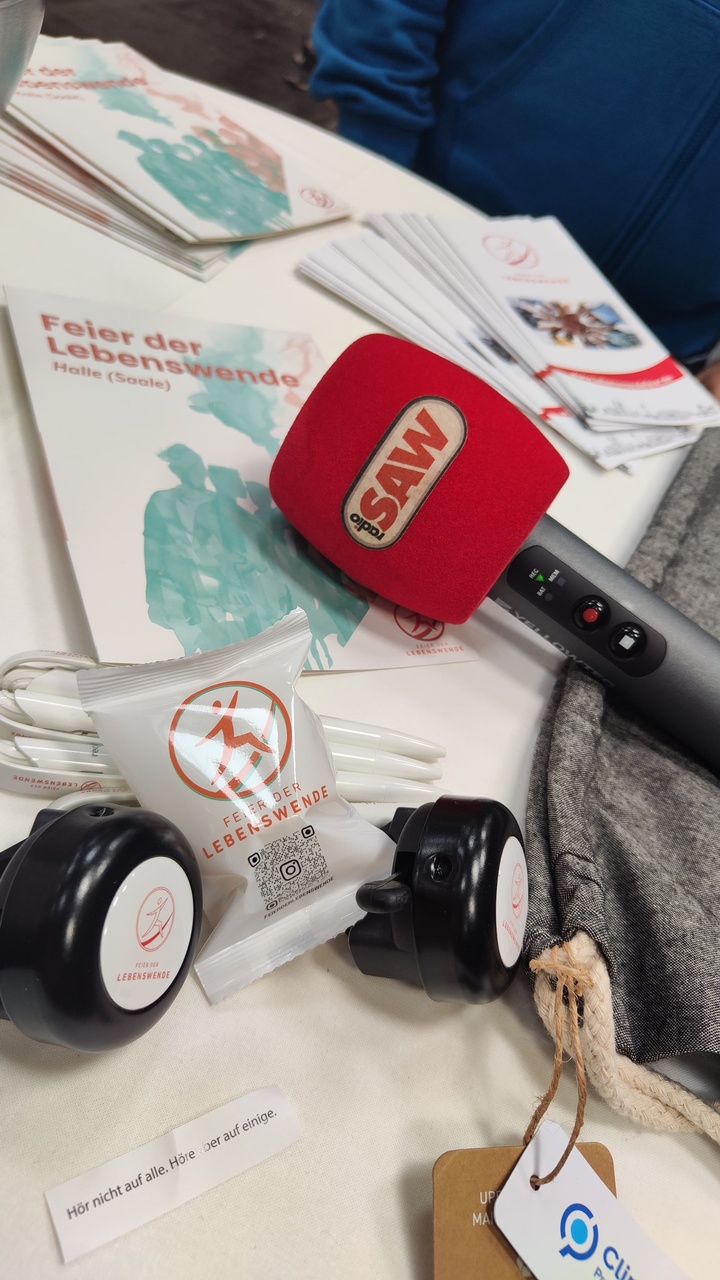 Nahaufnahme eines Stehtisches, Mikrofon von Radio SAW, Glückskeksverpackung, Fahrradklingel und Flyer liegen auf dem Tisch.