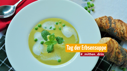 Ein weißer Porzellanteller gefüllt mit grünlicher Erbsensuppe. Daneben, auf dem Tisch liegen ein paar Stück Brot und ein Löffel