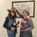 Zwei Frauen bei einer Hutübergabe im Kinder- und Jugendzentrum Don Bosco. Eine Frau trägt einen braunen Cowboyhut und hält mehrere Handpuppen, während die andere ihr den Hut aufsetzt und eine blau-silberne Dekoration in den Händen hält. Beide lächeln in d