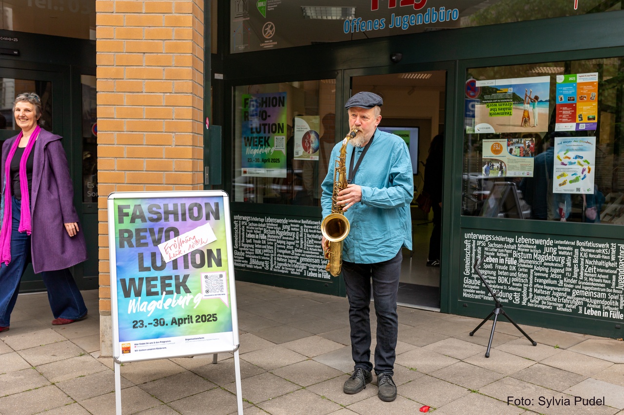 Mann spielt Saxophone vor einer offenen Tür. Neben ihn steht ein Aufsteller mit einem Plakat zur Fashon Revolution Week Magdeburg.