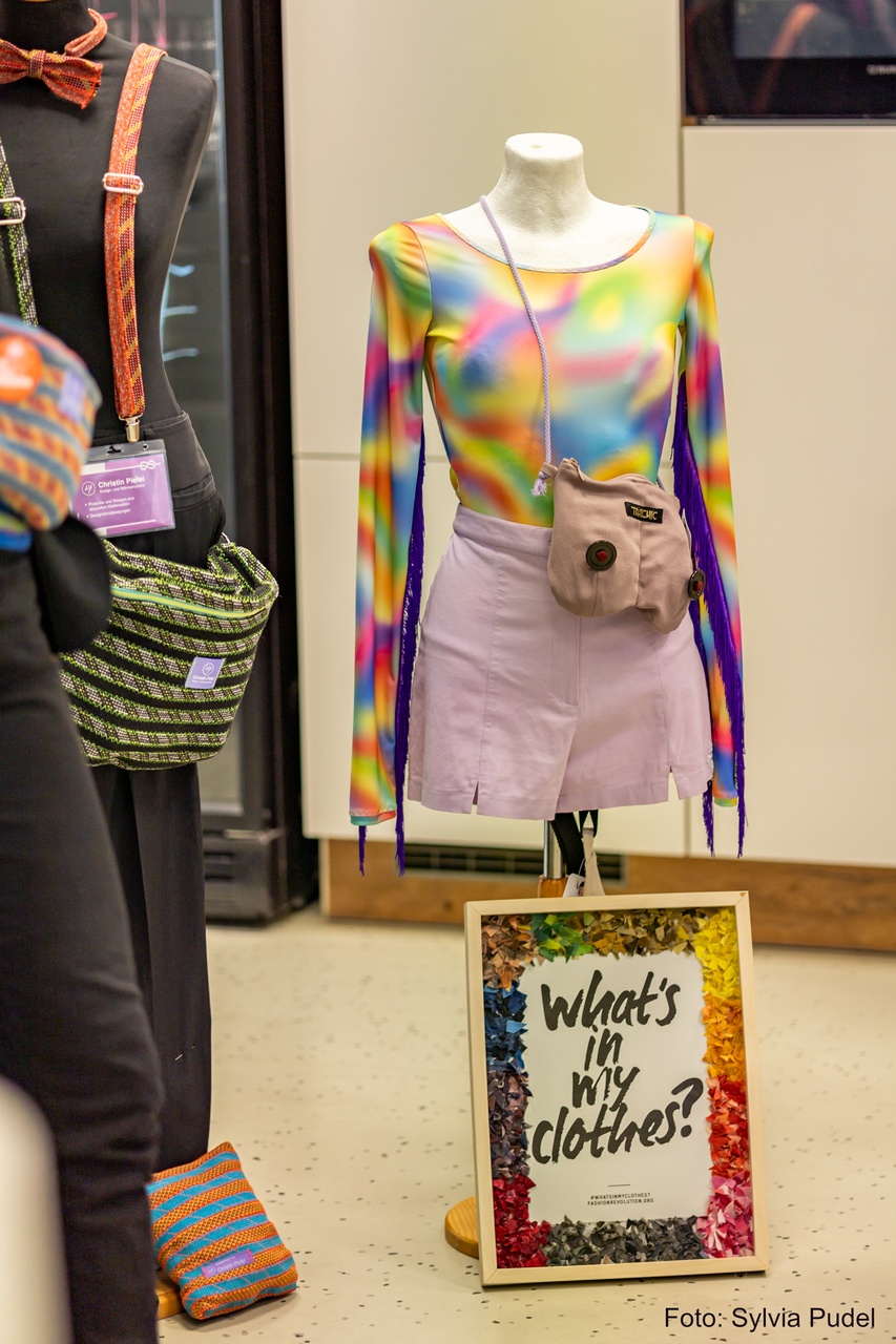 Eine Modepuppe mit einem regenbogenfarbenen Sportbody, und rosafarbenen Rock und einer Umhängetasche in Brustform. Davor steht ein Bilderrahmen mit die Worten: What´s in my clothes?