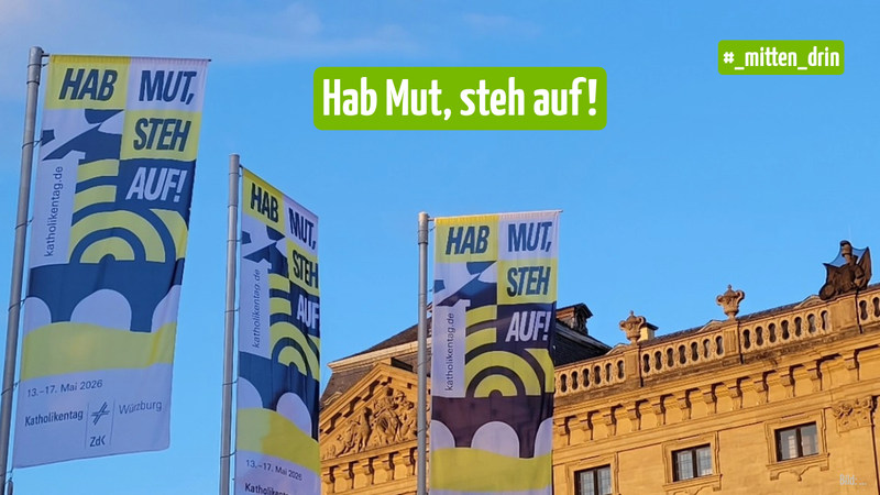 Fahnen vor einem großen Gebäude. Auf den Fahnen steht "hab Mut, steh auf!"
