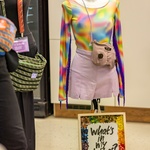 Eine Modepuppe mit einem regenbogenfarbenen Sportbody, und rosafarbenen Rock und einer Umhängetasche in Brustform. Davor steht ein Bilderrahmen mit die Worten: What´s in my clothes?