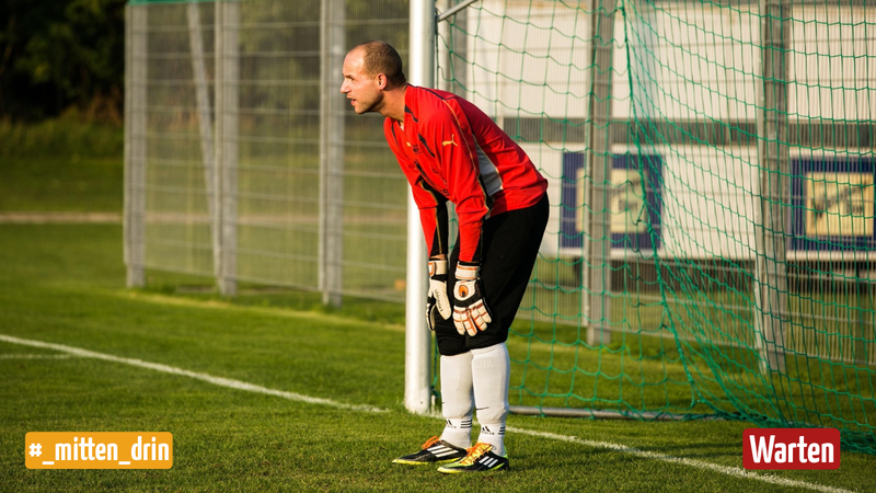 Ein Mann steht im Fußballtor. Er trägt ein rotes Trikot, eine schwarze Hose, Fußballschuhe und Torwarthandschuhe.