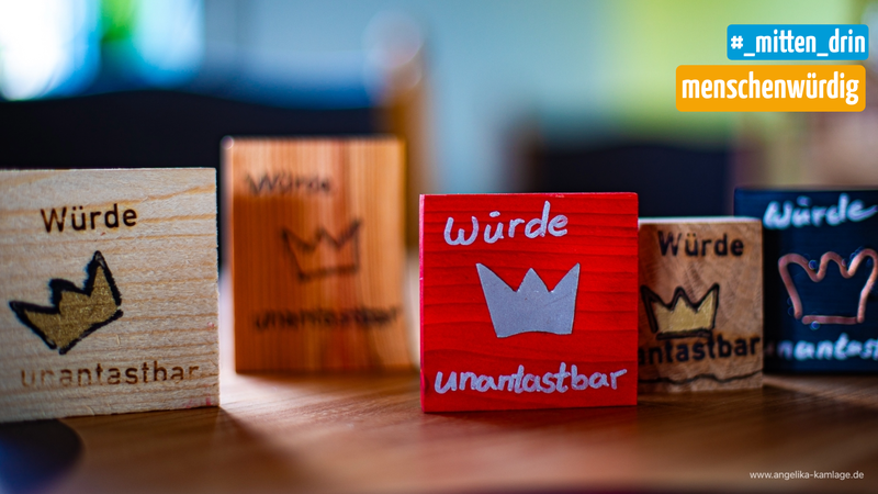 Mehrere bunte Holzklötze mit der Aufschrift „Würde unantastbar“ und kleinen Kronen stehen auf einem Tisch. Im Hintergrund ist unscharf das Logo mit den Worten „#_mitten_drin“ und „menschenwürdig“ zu sehen.
