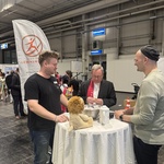 3 Männder am Stehtisch an einem Messestand. In der Mitte Bischof Bätzing. Auf dem Stehtisch steht ein Kuscheltier (Löwe) sowie eine Schüssel mit Keksen.