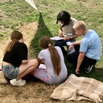 Zwei Mädchen, ein Junge und eine Frau sitzen bzw. hocken auf einer Wiese und sind von hinten und von der Seite zu sehen. Sie schauen auf ein Blatt Papier, welches das eine Mädchen und die Frau vor sich liegen haben.