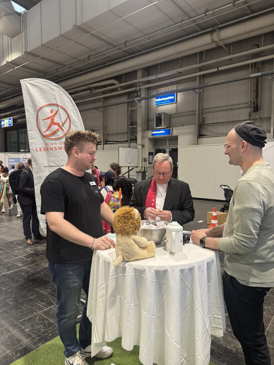 3 Männder am Stehtisch an einem Messestand. In der Mitte Bischof Bätzing. Auf dem Stehtisch steht ein Kuscheltier (Löwe) sowie eine Schüssel mit Keksen.