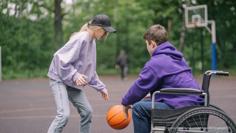Eine junge Frau (stehend) spielt Basketball mit einem jungen Mann, der im Rollstuhl sitzt