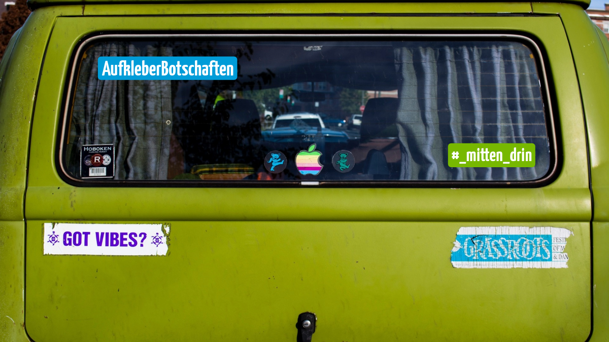 Heck eines grünen Vans mit Rückfenster und mehreren Aufklebern. Zu sehen sind bunte Symbole, der Schriftzug „AufkleberBotschaften“, der Hashtag „#_mitten_drin“ sowie Sticker mit Texten wie „GOT VIBES?“.
