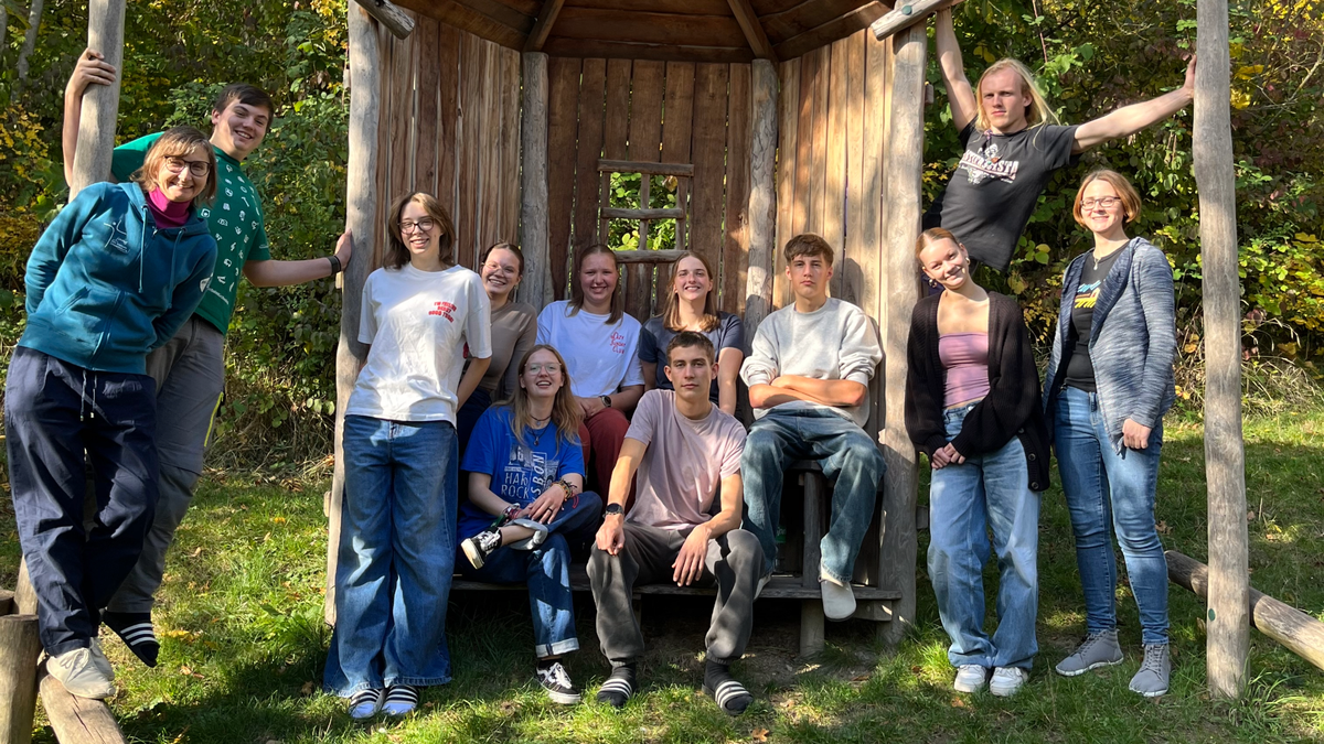 In einer kleinen Holzhütte und auch drumherum stehen 12 junge Menschen zu sehen.